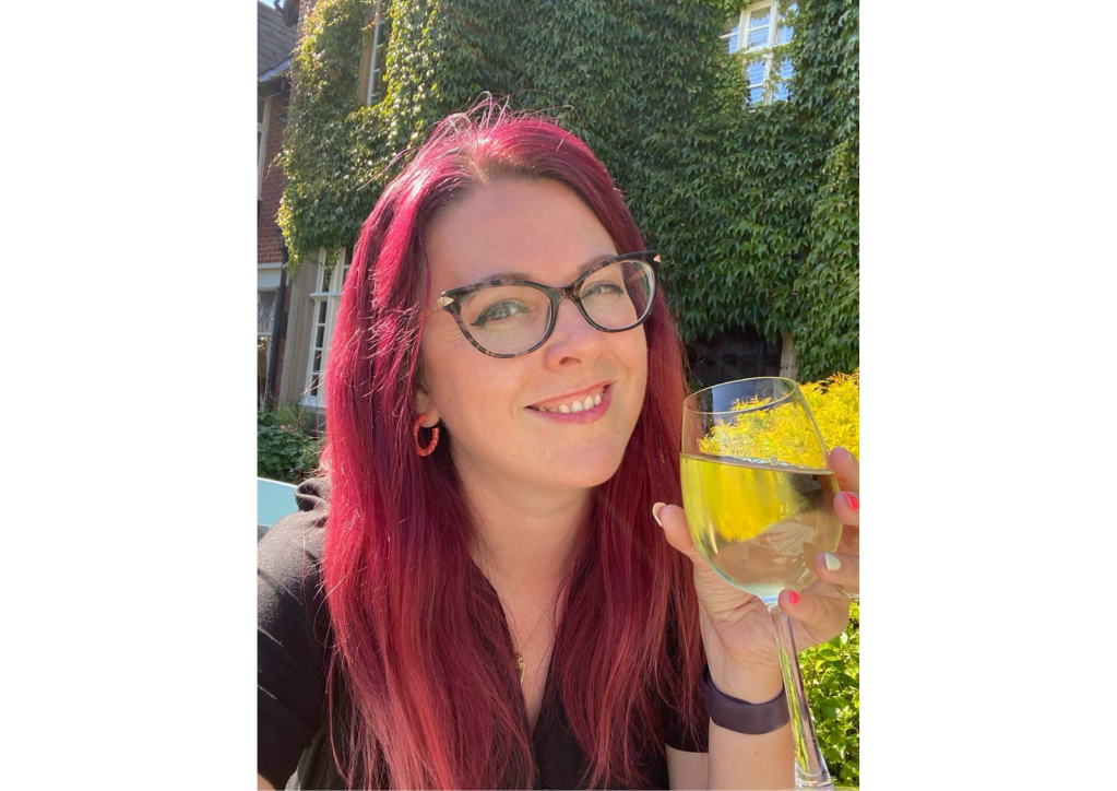 A white woman with bright red hair and glasses holding a glass and smiling